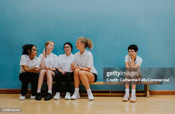 young teenagers sit on a bench in a school gymnasium, ignoring one of the other girls and cruelly excluding her - stereotype stockfoto's en -beelden