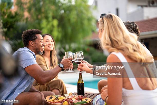 diverse friends toasting with wine and sharing food by the pool on a bright day - poolside stock pictures, royalty-free photos & images