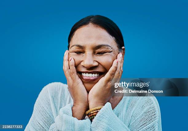joyful middle aged latina woman smiling with eyes closed and hands on cheeks blue background - self-love stock pictures, royalty-free photos & images
