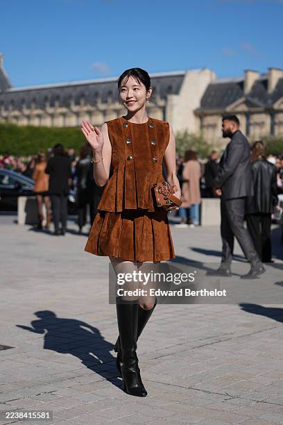 Yuka Kouri wears black hair pulled into a low bun with short curtain bangs, gold hoop earrings and gold rings and a gold bracelet, a brown leather...