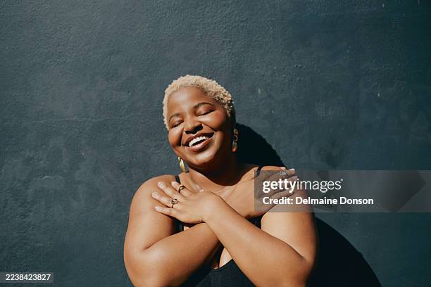 joyful black woman embracing herself with eyes closed smiling against dark background - body positive stock pictures, royalty-free photos & images