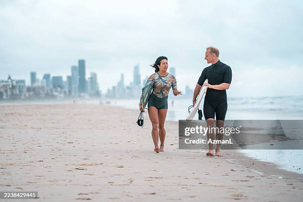 joyful mid adult surfing couple strolling on gold coast beach - barefoot stock pictures, royalty-free photos & images
