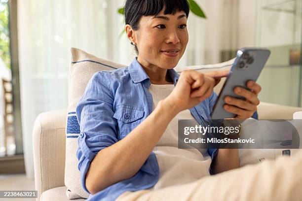 woman sitting comfortably while using a smartphone in a modern home setting - smirking stock pictures, royalty-free photos & images
