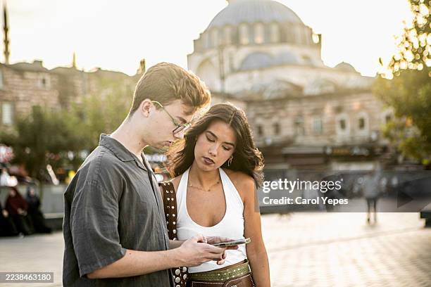 young couple smiling while looking at a smartphone in a square in istanbul - kabelloses-laden-von-gerät-zu-gerät stock-fotos und bilder