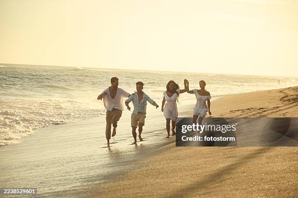 quattro amici corrono lungo una spiaggia illuminata dal sole al tramonto, condividendo la gioia in abiti bianchi - oceano indiano foto e immagini stock