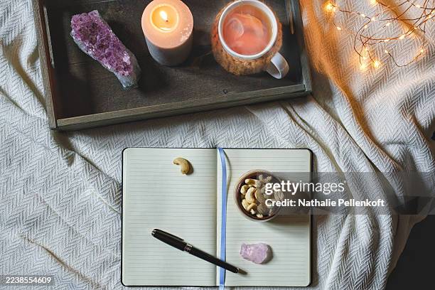 cozy evening scene with diary on bed, featuring amethyst crystal, cup of tea, candle and bowl of cashews - rose quartz stock pictures, royalty-free photos & images