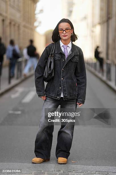 Rebecca Ragot-Montus is seen wearing rectangular glasses, gold hoop earrings, a purple checkered tie over a collared white shirt, a black denim...