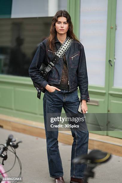 Airida Lopetaitė is seen wearing a dark denim cropped jacket, a sheer black top, dark denim straight-leg jeans, brown loafers, and carrying a black...