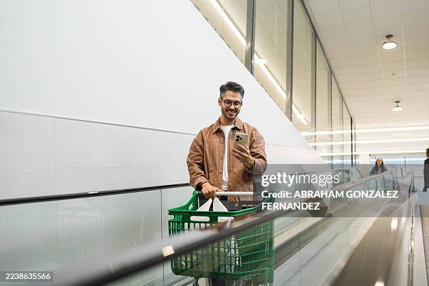 hombre adulto joven sonriendo mientras usa su teléfono móvil y empuja un carrito de compras en una acera inclinada en movimiento - hombres de mediana edad fotografías e imágenes de stock