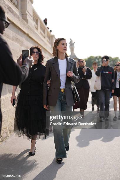 Rosamund Pike wears a side-parted medium-length light brown hairstyle, gold rings, a black leather shoulder bag with diamond-quilted stitching and a...