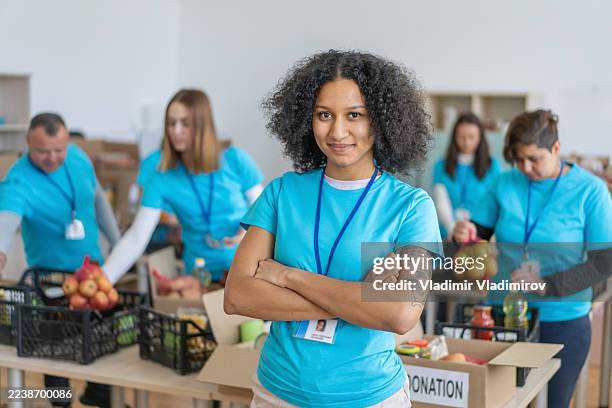 jeune bénévole souriant les bras croisés devant les dons de nourriture - altruisme photos et images de collection