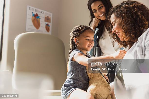 un médecin examine un enfant avec un ours en peluche alors que la mère sourit de manière rassurante lors d’un rendez-vous médical. - pédiatre photos et images de collection