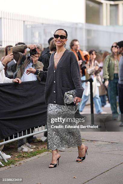 Sabrina Sato wears sleek pulled-back dark hair in a low bun, black cat-eye sunglasses, silver hoop earrings with stacked silver bracelets and a gold...