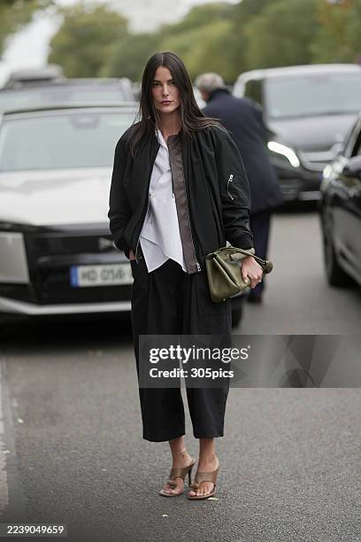 Leia Sfez is seen wearing a black oversized bomber jacket layered over a white button-up shirt, loose black trousers, brown heeled sandals, and...