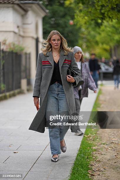 Guest wears loose wavy blonde hair parted in the center, a gold necklace, a handheld black leather clutch bag, a long grey wool coat with red and...