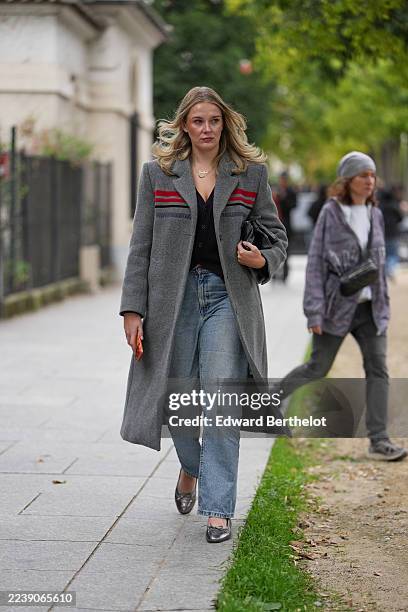 Guest wears loose wavy blonde hair parted in the center, a gold necklace, a handheld black leather clutch bag, a long grey wool coat with red and...
