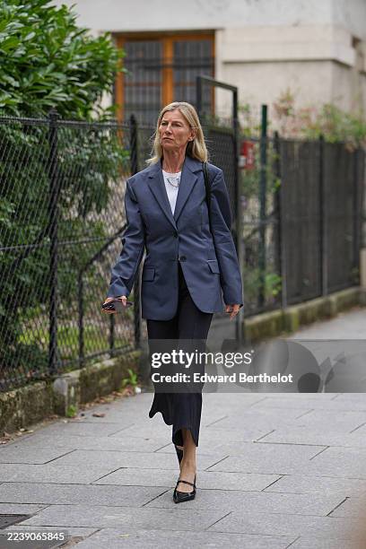 Guest wears shoulder-length straight blonde hair with a center part, a silver chain necklace, a black leather shoulder bag, a white ribbed crewneck...