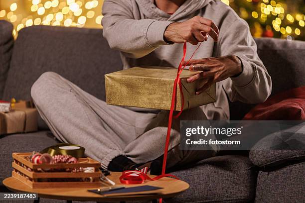 eine person in einem grauen kapuzenpullover wickelt zu weihnachten ein goldenes geschenk mit roter schleife auf ein gemütliches wohnzimmersofa - gift lounge stock-fotos und bilder