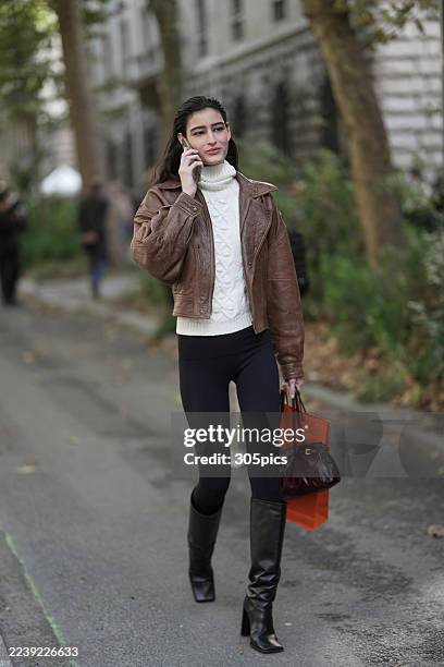 Luiza Perote is seen wearing a brown leather jacket, a white cable-knit turtleneck sweater, black leggings, black heeled knee-high boots, and...