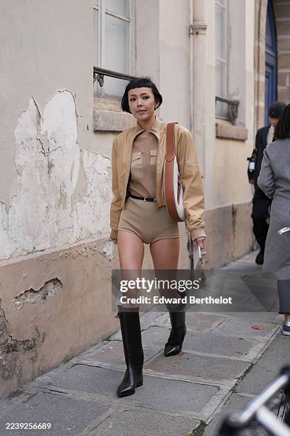 Kiwi Lee Han wears a short straight black bob hairstyle with blunt micro bangs, silver hoop and stud earrings, a brown leather shoulder bag with...