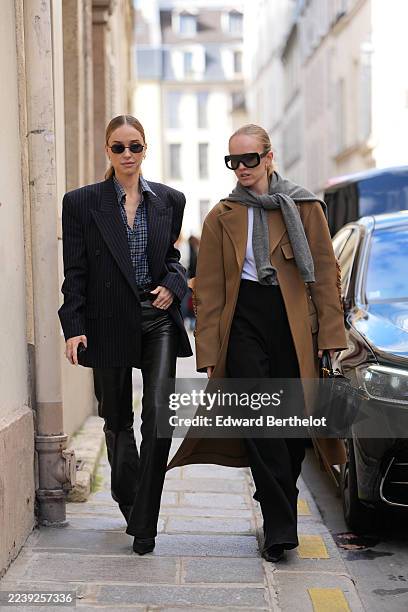 Guest wears a pulled-back low ponytail of light brown hair with a center part, small oval black sunglasses, gold hoop earrings, a brown leather...