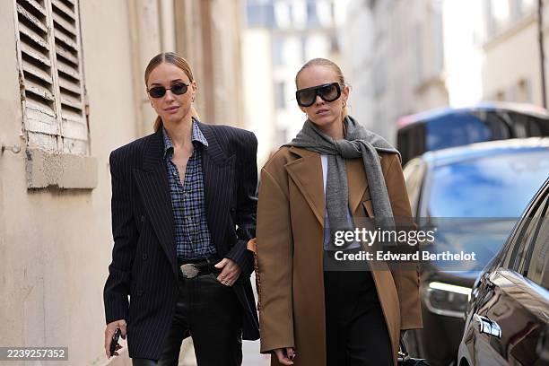 Guest wears a pulled-back low ponytail of light brown hair with a center part, small oval black sunglasses, gold hoop earrings, a brown leather...