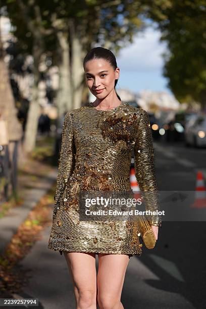 Guest wears a sleek low ponytail hairstyle in dark brown with a center part, small stud earrings, a gold metallic round clutch bag, a long-sleeve...