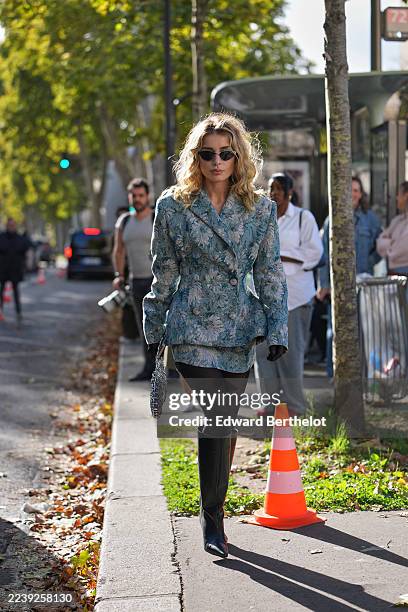 Guest wears wavy shoulder-length blonde hair with a middle part, cat-eye black sunglasses, a silver chain-link handheld bag, black leather gloves, a...