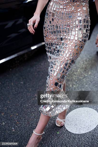 Guest wears a nude sheer maxi dress embellished with mirror-like panels, paired with strappy white heels, bold earrings, and small black sunglasses...