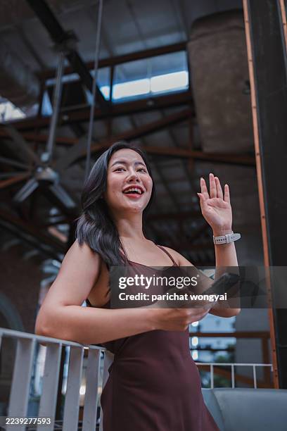 young adult asian woman in a casual dress smiling and waving, holding a smartphone in an urban setting - medium shot stock pictures, royalty-free photos & images