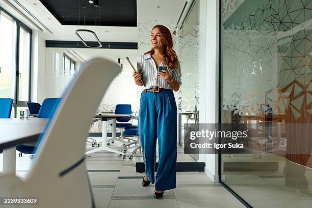 businesswoman walking through modern office carrying clipboard and phone - trousers stock pictures, royalty-free photos & images