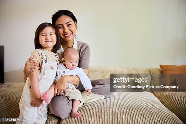 portrait of a joyful mother sitting with her two children in her arms on a sofa in bright living room, smiling at camera - family with two children stock pictures, royalty-free photos & images