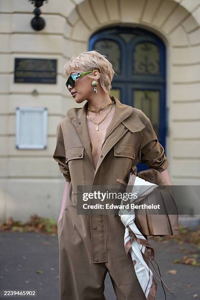 Michelle Dee wears a short layered blonde pixie haircut, rectangular green-tinted sunglasses, layered gold chain necklaces and gold hoop earrings, a...