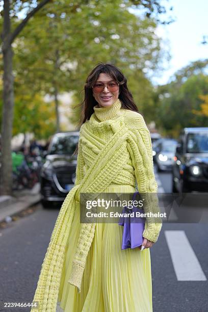 Guest wears long wavy brown hair with a center part, brown-tinted oversized sunglasses, a purple leather clutch bag, a yellow chunky cable-knit...