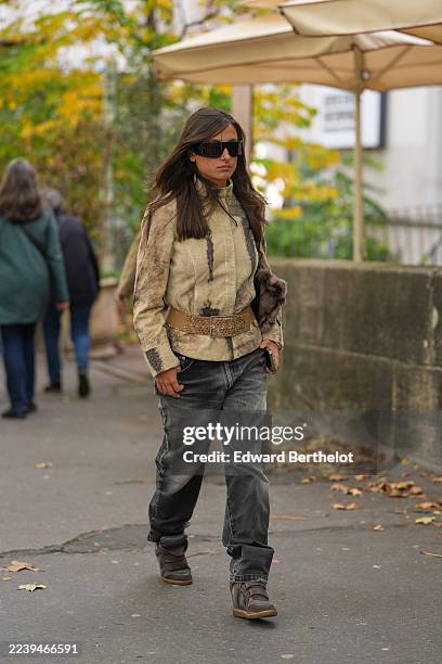 Guest wears long straight dark brown hair with a slight center part, large rectangular black futurist sunglasses, a brown fur clutch bag, a beige...