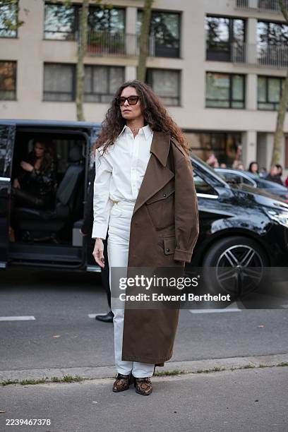 Guest wears long dark brown curly hair parted in the center, rectangular tortoiseshell brown sunglasses, a thin gold chain necklace, a white...