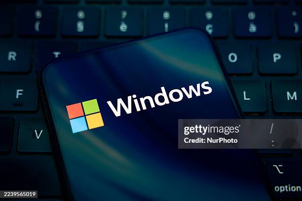 The Windows logo appears on the screen of a smartphone placed on a computer keyboard, which reflects an abstract illustration in blue colors....