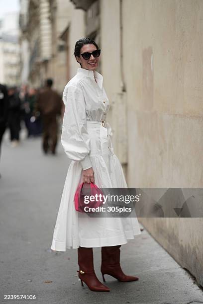 Sally Holmes is seen wearing dark hair styled back, oversized black sunglasses, layered necklaces, a long-sleeve white belted midi shirt dress with...