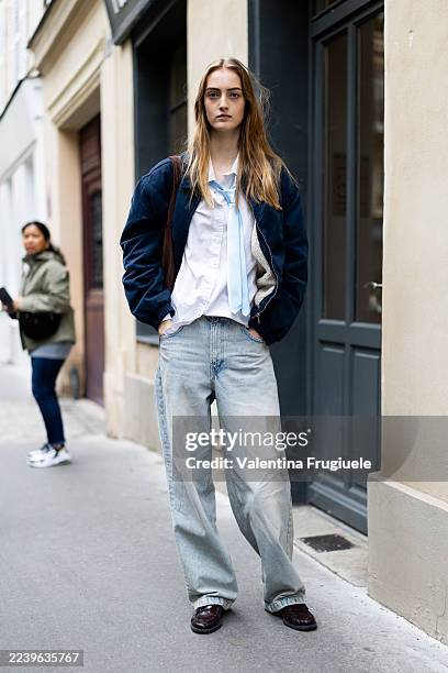 Model wears a navy bomber jacket over a white shirt with a light blue tie, light wash baggy jeans, and burgundy loafers, carrying a brown tote bag...