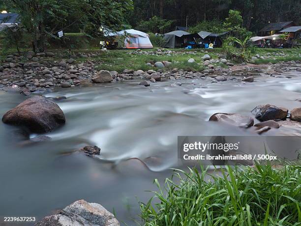 long exposure shot of a river flowing over rocks with camping tents set up on the bank surrounded by lush trees - estado de selangor fotografías e imágenes de stock