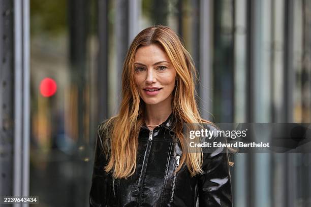 Phoebe Tonkin wears long wavy light brown hair with blonde highlights, middle part, a black patent varnished leather zip-front jacket with collar and...