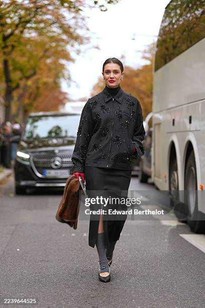 Kate Sardarov wears hair pulled into a low bun, medium brown hair with a center part, white pearl stud earrings, a brown fur clutch bag, a black...