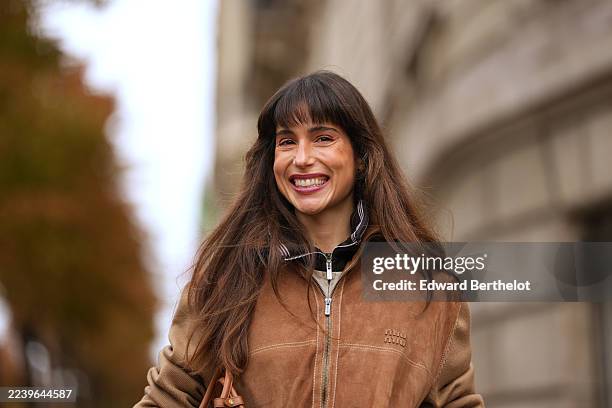 Belen Hostalet wears long straight brown hair with blunt bangs, a camel suede bomber jacket with a ribbed black-and-white striped collar and ribbed...