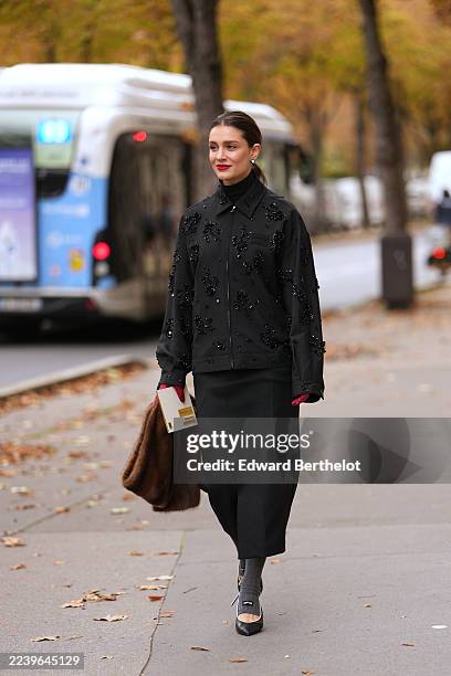 Kate Sardarov wears hair pulled into a low bun, medium brown hair with a center part, white pearl stud earrings, a brown fur clutch bag, a black...