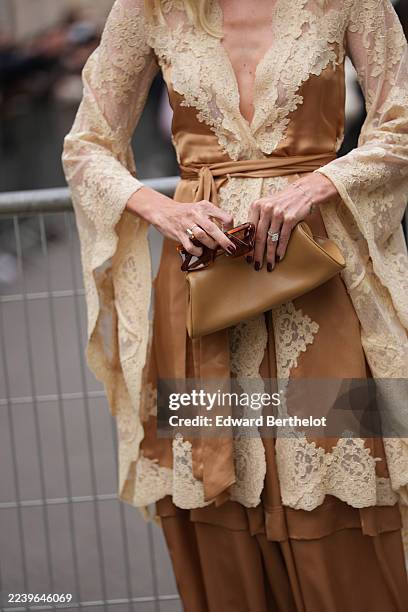 Devon Windsor wears a metallic gold leather clutch bag, a champagne-gold satin wrap top with a deep V neckline and cream lace trim and sheer cream...