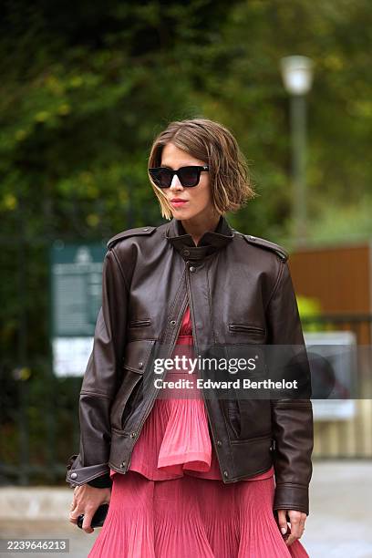 Monica Ainley wears a chin-length light brown bob with a soft center part, black rectangular sunglasses, a dark brown leather jacket with epaulets, a...