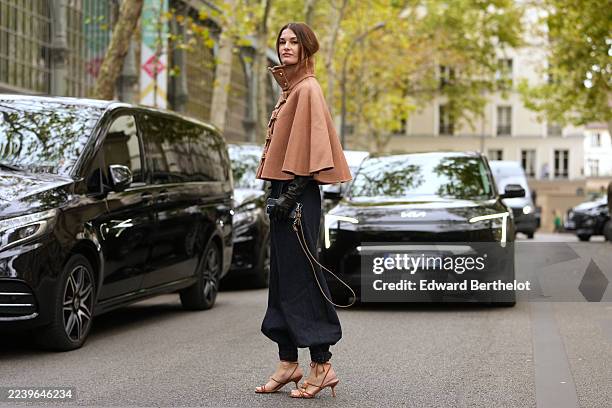 Ophelie Guillermand wears medium-brown hair pulled back into a low bun with a center part, long gold drop earrings, a camel wool cape jacket with a...