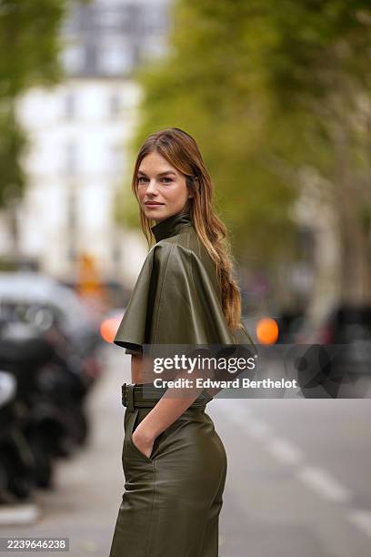 Nadine Leopold wears long straight light brown hair with blonde highlights, center part, olive green leather cropped turtleneck top with wide short...