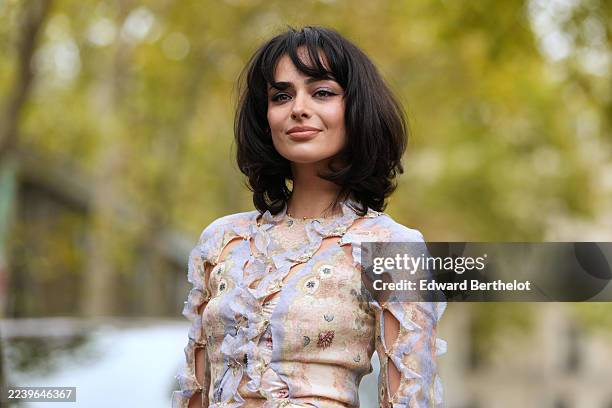 Aubri Ibrag wears a chin-length dark brown bob with blunt bangs, a long-sleeve sheer pastel floral paisley printed chiffon maxi dress with cascading...