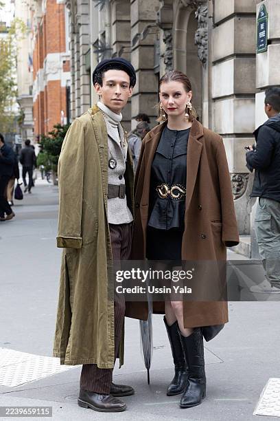 Guest wears a long olive green trench coat layered over a beige turtleneck and striped brown trousers, paired with brown shoes, while another guest...